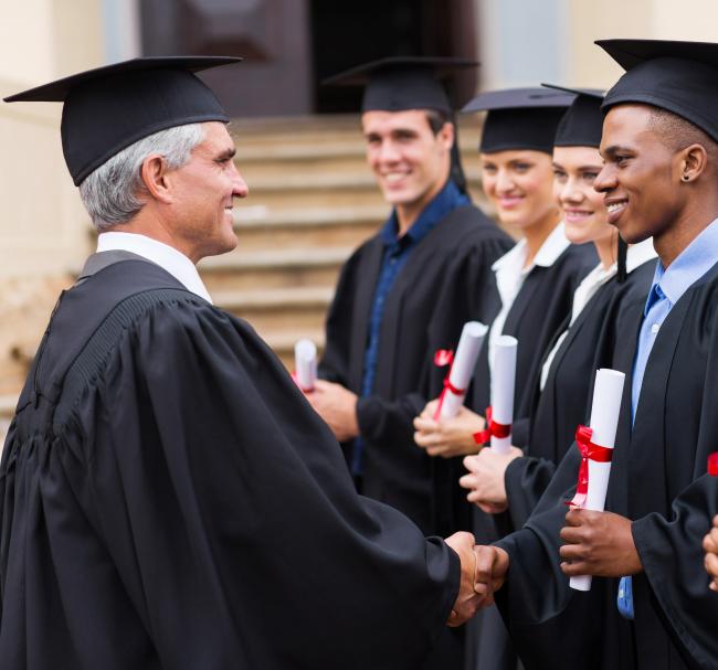 University president in cap and gown shaking hands of graduates holding degrees